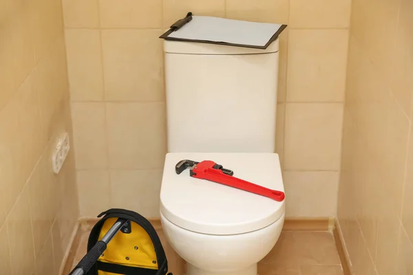 Pipe wrench and clipboard on toilet indoors — Stock Photo, Image