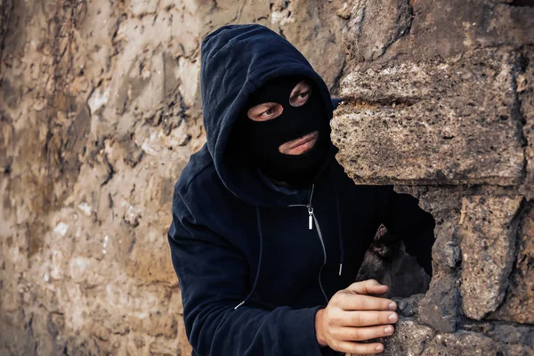 Homem mascarado atrás da parede de pedra a ver alguém ao ar livre. Actividade criminosa — Fotografia de Stock