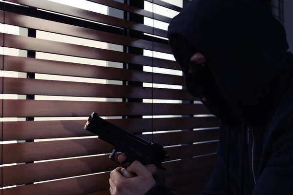 Maskerad man med pistol spionerar genom fönsterpersienner inomhus. Farliga kriminella — Stockfoto