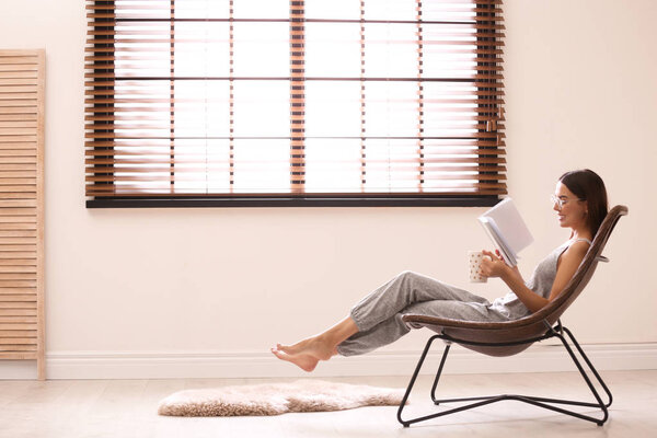 Young woman reading book near window with blinds at home. Space for text
