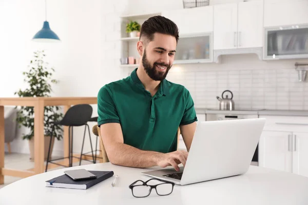 Handsome young man working with laptop at table in kitchen — Stock Photo, Image