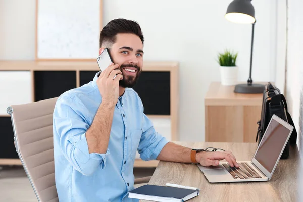 Knappe jonge man praten op telefoon tijdens het werken aan tafel met laptop in thuiskantoor — Stockfoto