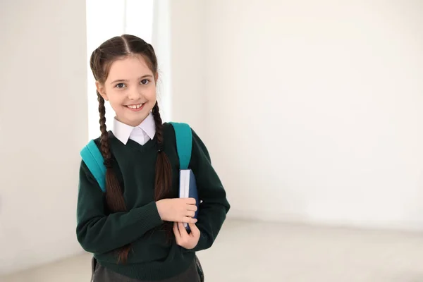 Retrato de menina bonito em uniforme escolar com mochila e livro dentro de casa. Espaço para texto — Fotografia de Stock