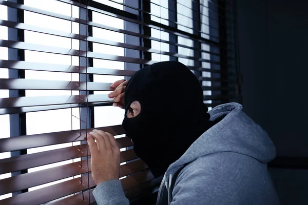 Маска людина шпигує через віконні жалюзі в приміщенні. Кримінальне правопорушення — стокове фото