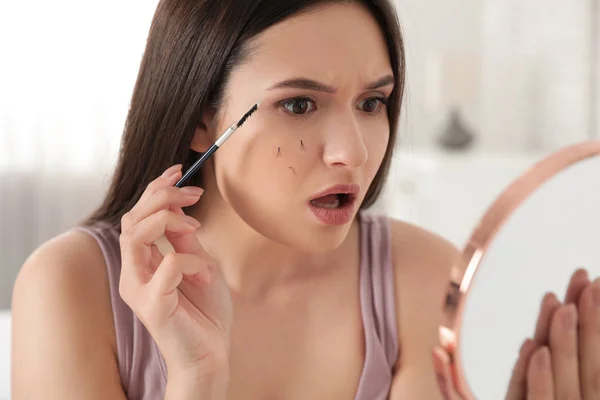Beautiful woman with fallen eyelashes and cosmetic brush looking into mirror indoors — Stock Photo, Image