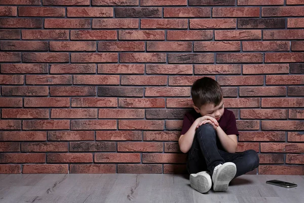 Niño triste con teléfono móvil sentado en el suelo cerca de la pared de ladrillo. Espacio para texto —  Fotos de Stock