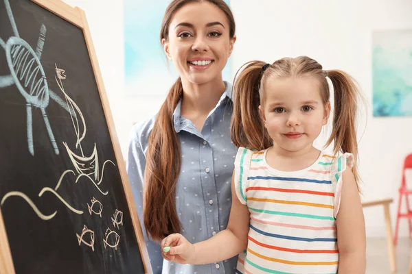 Kindergarten teacher and little child near chalkboard indoors. Learning and playing