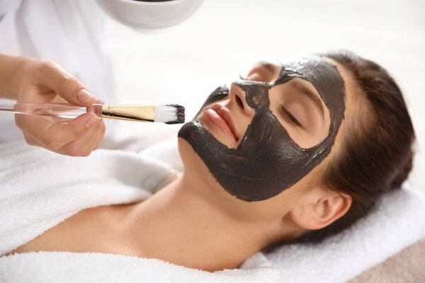 Cosmetologist applying black mask onto woman's face in spa salon