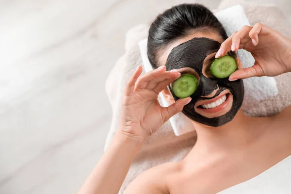 Vacker kvinna med svart mask på ansiktet och gurka skivor koppla av i Spa-salongen, uppifrån. Utrymme för text — Stockfoto