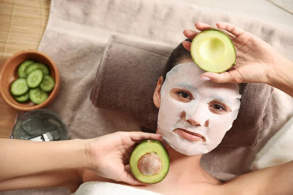 Beautiful woman with cotton mask sheet on face and cut avocado relaxing in spa salon, top view — Stock Photo, Image