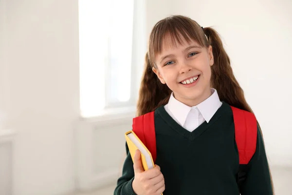 Retrato de menina bonito em uniforme escolar com mochila e livro dentro de casa. Espaço para texto — Fotografia de Stock