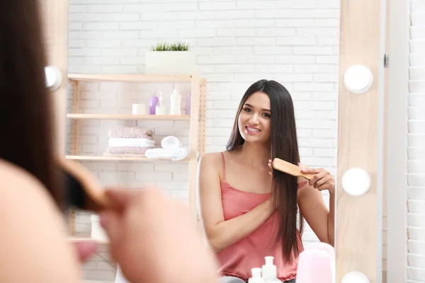 Belle jeune femme avec brosse à cheveux regardant dans le miroir dans la salle de bain — Photo