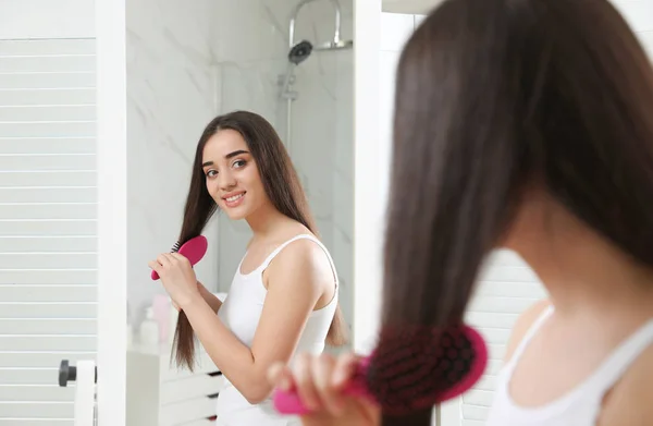Mulher bonita com escova de cabelo olhando para o espelho no banheiro — Fotografia de Stock