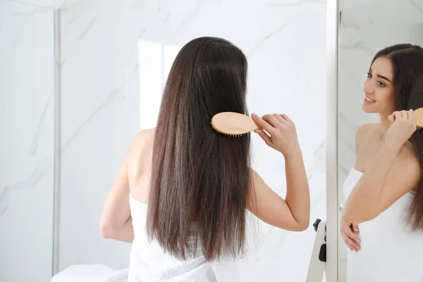 Beautiful young woman with hair brush looking into mirror in bathroom — Stock Photo, Image