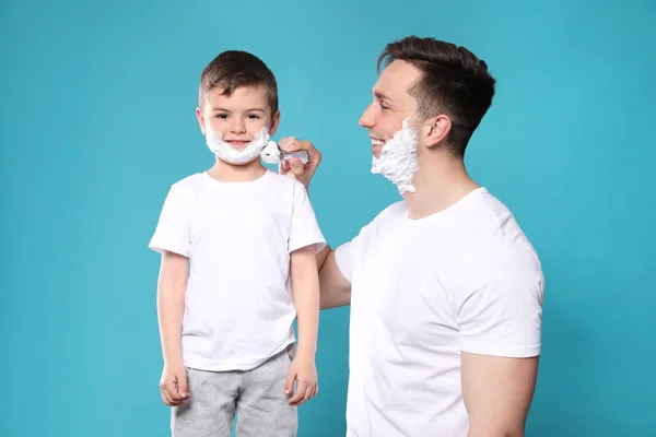 Papá aplicando espuma de afeitar en la cara del hijo contra el fondo de color — Foto de Stock