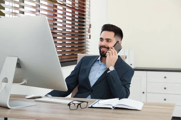 Knappe zakenman praat op telefoon tijdens het werken met computer aan tafel in Office — Stockfoto