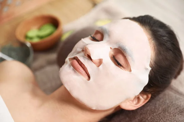 Mulher bonita com folha de máscara de algodão no rosto relaxante no salão de spa, close-up — Fotografia de Stock