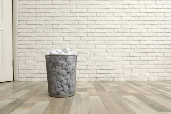 Metal bin with crumpled paper on floor against brick wall, space for text