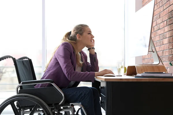 Kobieta na wózku inwalidzkim pracuje z komputerem przy stole w pomieszczeniu — Zdjęcie stockowe