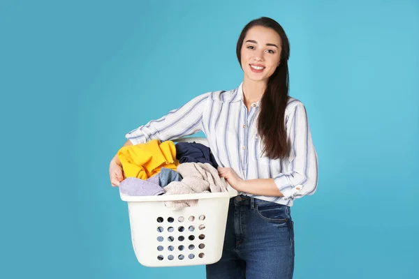 Happy young woman holding basket with laundry on color background
