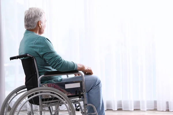 Uomo anziano seduto in sedia a rotelle vicino alla finestra a casa — Foto Stock