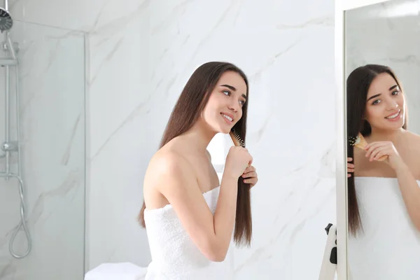 Hermosa mujer joven con cepillo de pelo mirando en el espejo en el baño — Foto de Stock