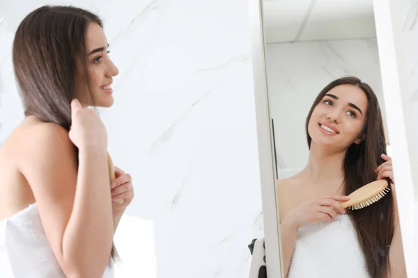 Красивая молодая женщина с расческой, смотрящая в зеркало в ванной комнате — стоковое фото