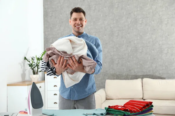 Mężczyzna trzyma stos ubrań w pobliżu deski do prasowania w domu. Miejsce na tekst — Zdjęcie stockowe