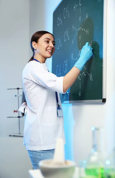 Cientista feminina escrevendo fórmula química em quadro-negro dentro de casa — Fotografia de Stock