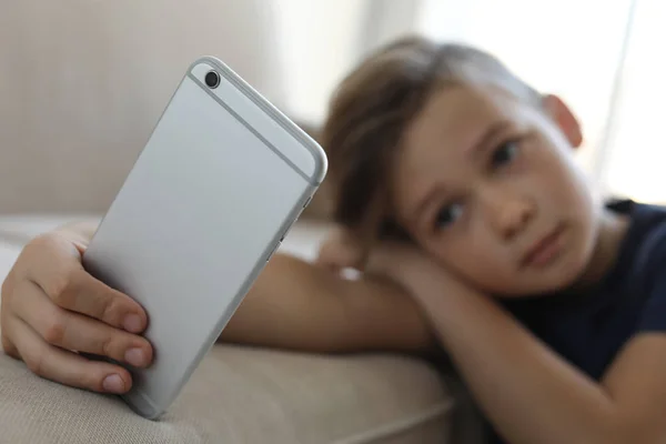 Сумна маленька дитина зі смартфоном, що спирається на диван у кімнаті. Небезпека Інтернету — стокове фото
