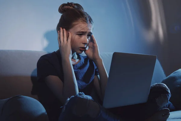 Menina adolescente chocada com laptop no sofá no quarto escuro. Perigo da internet — Fotografia de Stock