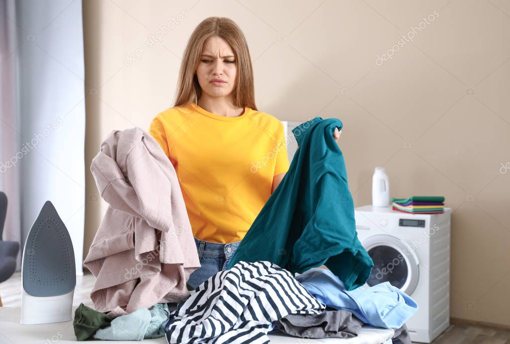 Emotional woman near board with iron and pile of clothes in bathroom