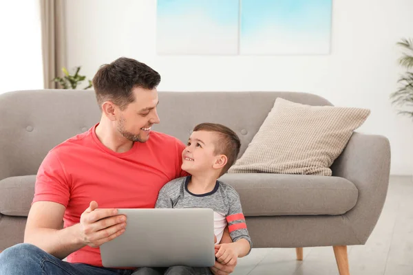 Мальчик и его отец с ноутбуком сидят рядом с диваном на полу. Семейное время — стоковое фото