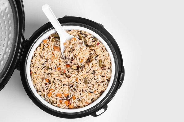 Delicious rice with vegetables and spoon in modern multi cooker on white background, top view