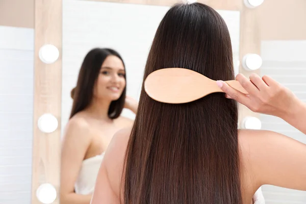 Hermosa mujer joven con cepillo de pelo mirando en el espejo en el baño — Foto de Stock