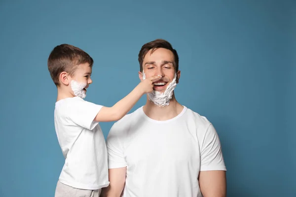 Papá y su pequeño hijo se divierten con espuma de afeitar en las caras contra el fondo de color — Foto de Stock
