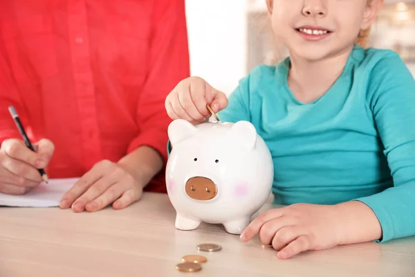 Uma menina a pôr moedas no mealheiro e a mãe à mesa, de perto. Poupar dinheiro — Fotografia de Stock