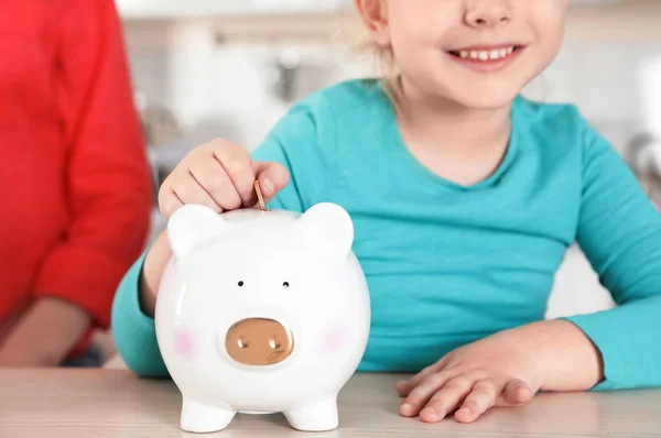 Schattig meisje zet munt in Piggy Bank aan tafel, close-up. Geld besparen — Stockfoto