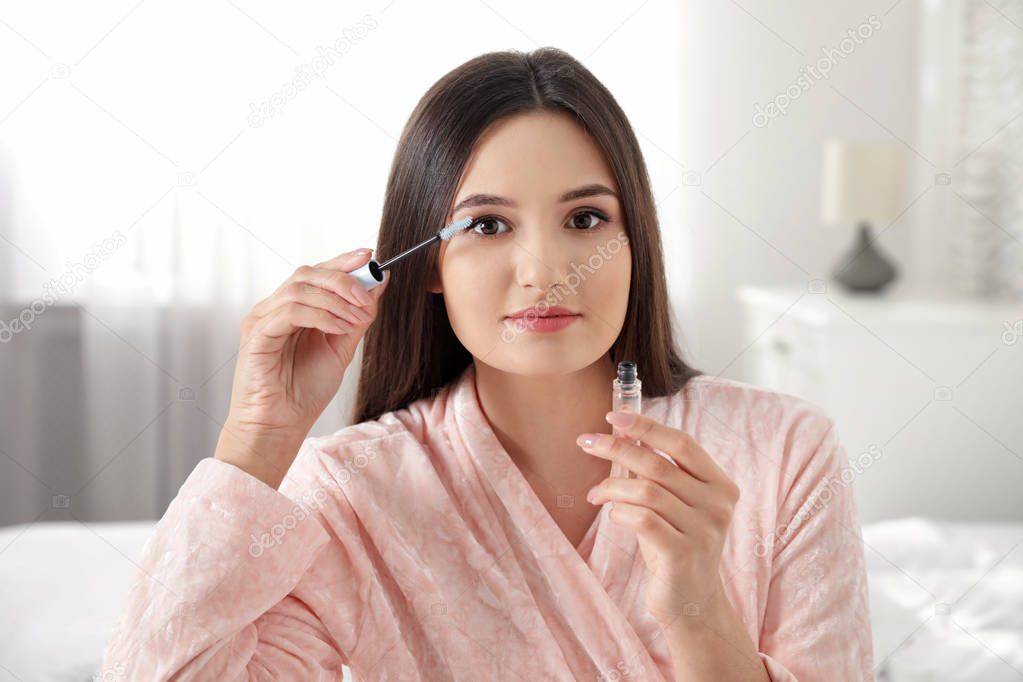 Beautiful woman applying oil onto her eyelashes indoors