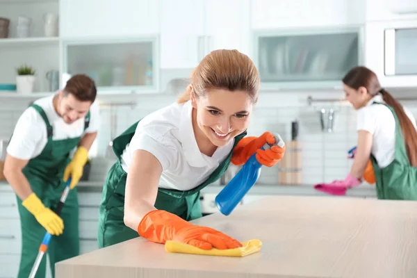 Mulher usando pano e pulverizador para limpeza de mesa com colegas na cozinha — Fotografia de Stock