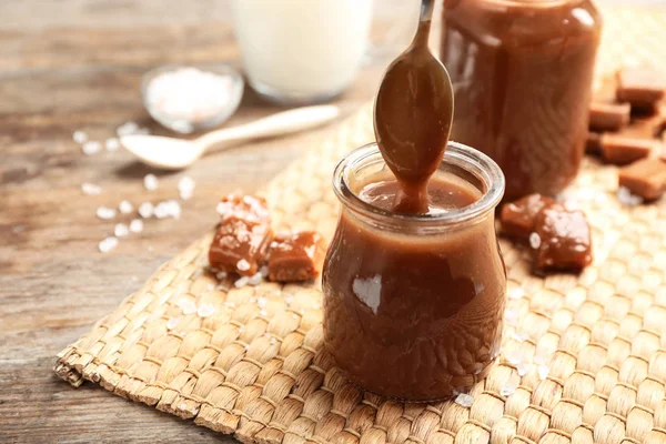 Spoon of caramel sauce above jar on wooden table. Space for text — Stock Photo, Image