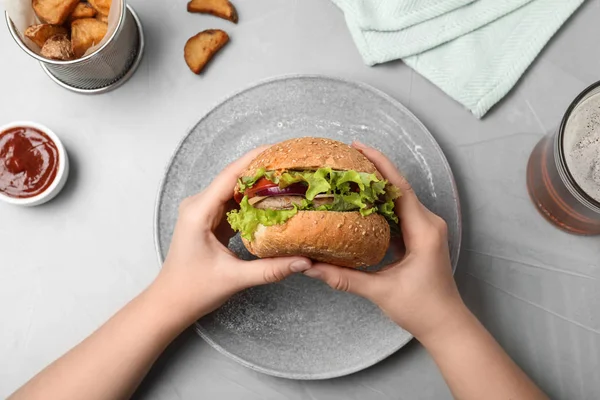 Mulher segurando saboroso hambúrguer sobre placa na mesa, vista superior — Fotografia de Stock
