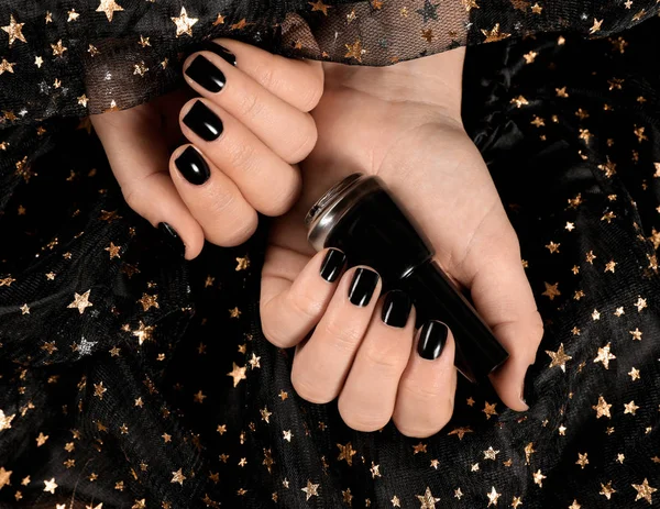 Woman with black manicure holding nail polish bottle over dark fabric, top view — Stok fotoğraf