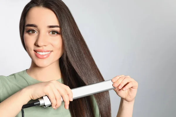 Mulher feliz usando ferro de cabelo no fundo cinza. Espaço para texto — Fotografia de Stock