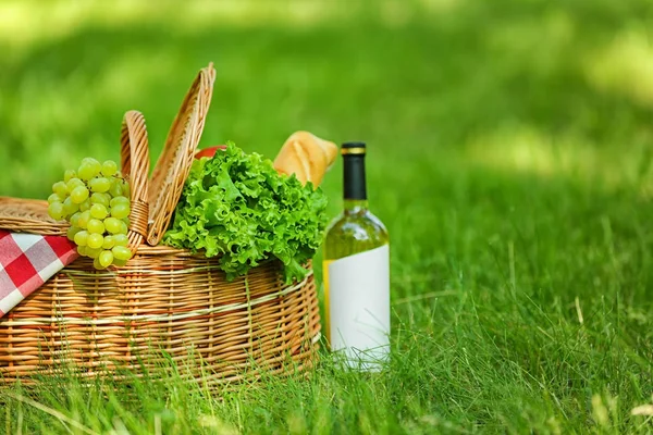 Плетеная корзина с одеялом, вином и едой на зеленой траве в парке, место для текста. Летний пикник — стоковое фото