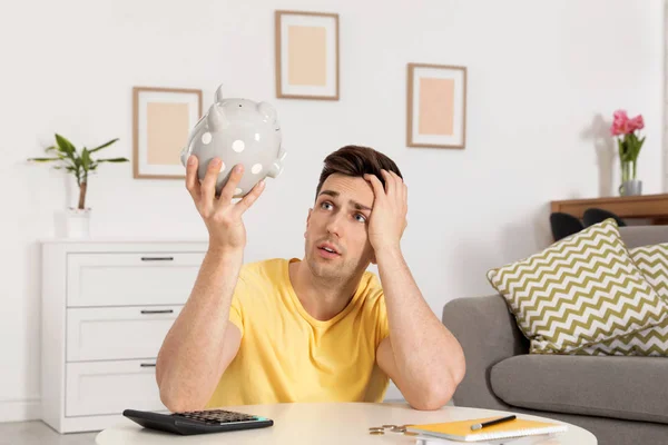 Sad man looking for money in piggy bank at table indoors