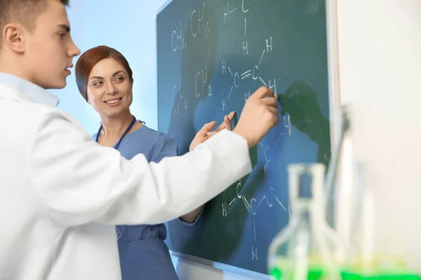 Scientists writing chemical formulas on chalkboard indoors — Stock Photo, Image