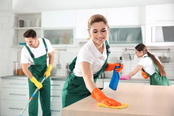 Mulher usando pano e pulverizador para limpeza de mesa com colegas na cozinha — Fotografia de Stock