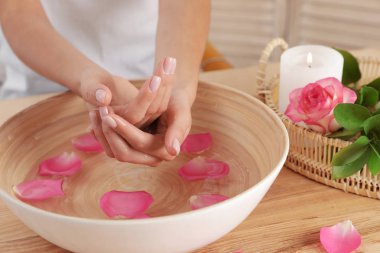Woman soaking her hands in bowl with water and petals on table, closeup. Spa treatment