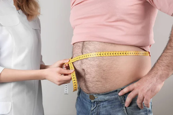 Doctor measuring overweight man's waist on color background, closeup